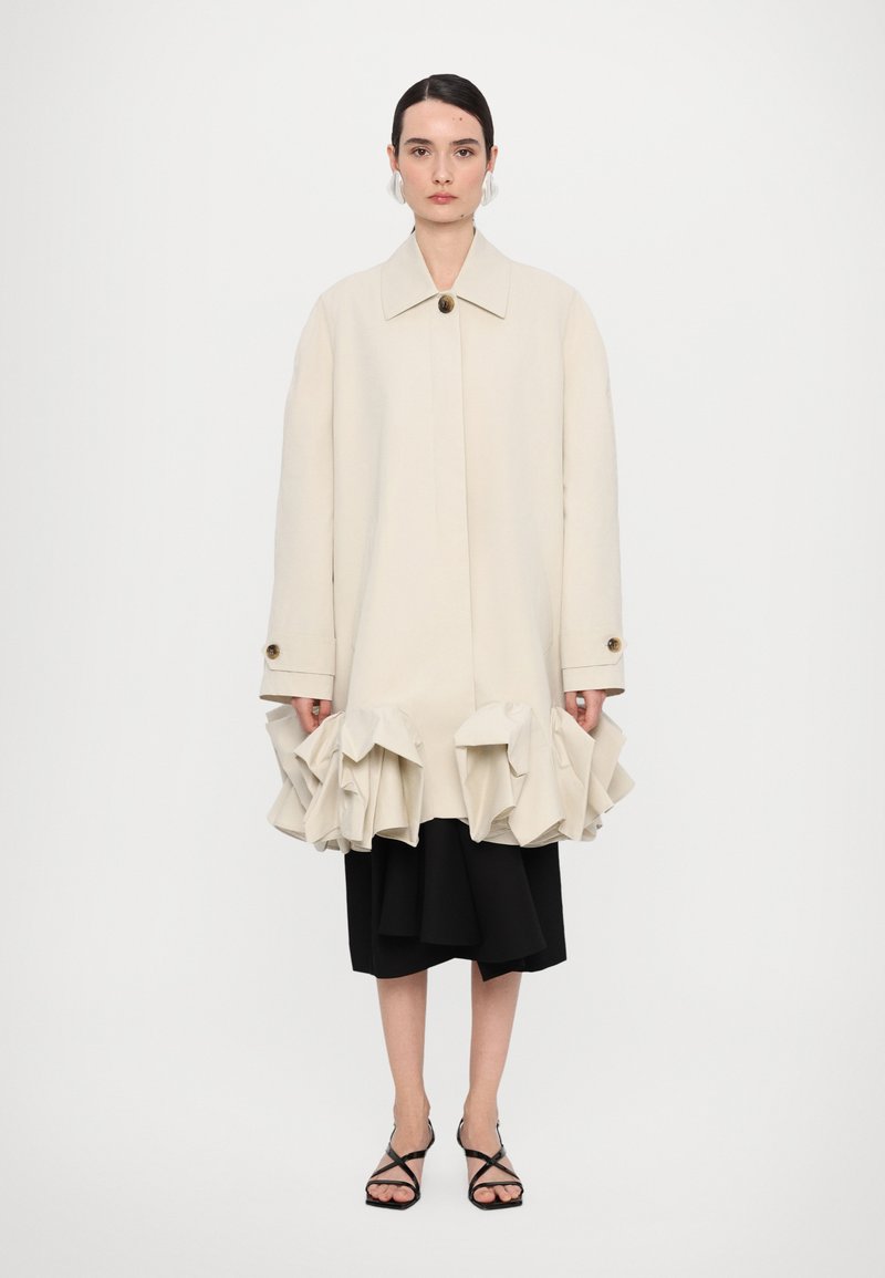 Beige tailored coat with a large ruffled hem, single button closure, and oversized fit, worn over a black layered skirt and strappy sandals.
