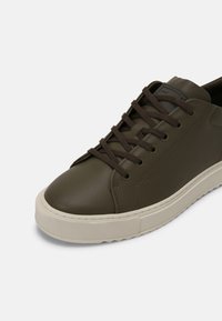 Sneakers z oliwkowej skóry, z okrągłym noskiem, brązowymi sznurowadłami, białą gumową podeszwą i delikatnymi detalami przeszycia. Minimalistyczny design z gładką teksturą.