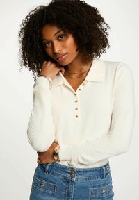 Polo shirt a maniche lunghe color crema chiaro con colletto e sette dettagli di bottoni dorati, abbinato a jeans di denim blu e accessori dorati.