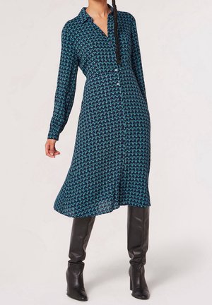 Kobieta w niebieskiej sukience midi z długim rękawem i wzorem, zapinanej na guziki, zestawiona z czarnymi, sięgającymi kolan, eleganckimi botkami na obcasie.