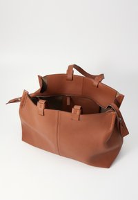 Sac fourre-tout en cuir marron avec un dessus ouvert, des poignées solides et un compartiment zippé. Texture lisse et design minimaliste.