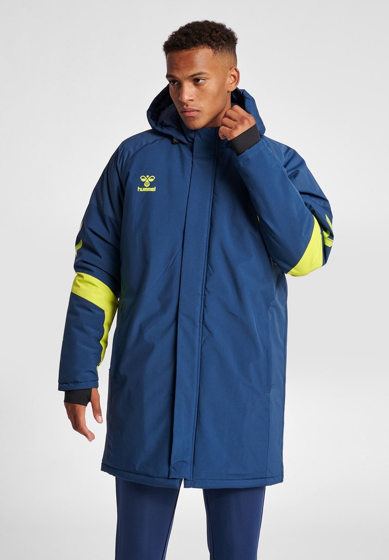 Young man adjusting collar wearing long blue winter coat with yellow accents and hood, standing against plain background.