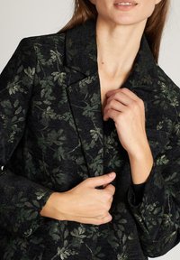 Mujer vestida con un blazer oscuro de patrón de hojas verdes, ajustando la solapa con ambas manos, mostrando el cuello y la parte inferior de la cara.
