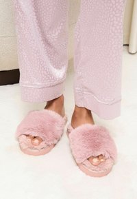 Papuci pufoși roz cu un upper din blană moale, cu vârf deschis și talpă din cauciuc, asortați cu pantaloni de pijama cu un model roz deschis.