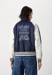 Sötétkék bomberdzseki fehér ujjakkal, csíkozott mandzsettákkal és hímzett "Scotch & Soda" logóval, amely épületmintát tartalmaz a hátsó részen.