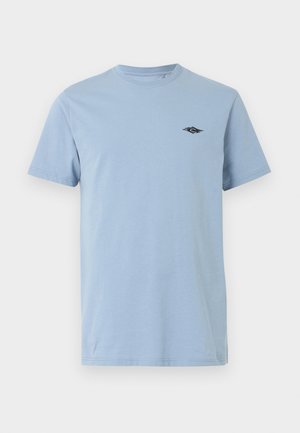 Giovane uomo con capelli scuri e ricci, che indossa una maglietta azzurro chiaro a maniche corte e pantaloni beige, in piedi davanti a uno sfondo semplice.