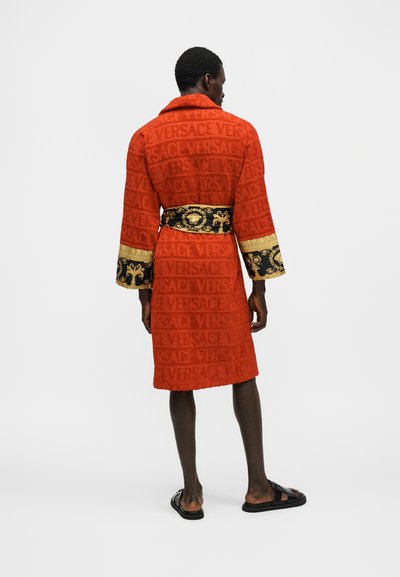 Άνδρας όρθιος με την πλάτη ορατή, φοράει κόκκινο ρόμπα Versace με ζώνη και μανσέτες σε μαύρο και χρυσό σχέδιο, σε συνδυασμό με μαύρα σανδάλια slide.