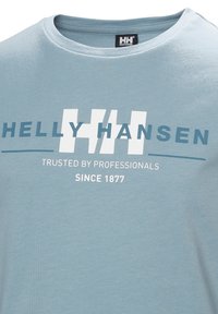 Camiseta de algodón azul claro con el texto del logo "Helly Hansen" en blanco y azul oscuro, con "Confiado por Profesionales" y "Desde 1877" impresos abajo.