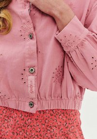 Różowa krótkia kurtka z haftowanymi detalami i srebrnymi guzikami, z elastycznymi mankietami. Nosi się ją na wzorzystą spódnicę w kwiaty w koordynujących kolorach.