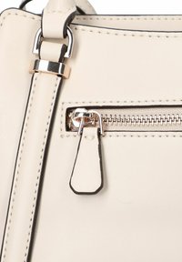 Détail d'un sac à main en cuir beige avec une poche zippée dotée d'une fermeture éclair métallique et d'un tirette, rehaussé de coutures et d'accessoires noirs.