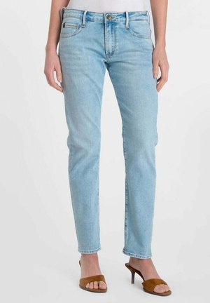 Jeans azzurri a gamba dritta indossati da una persona in piedi, abbinati a sandali con tacco marroni aperti sulla punta e una maglietta bianca.