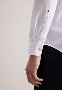 Mano maschile accanto al torso che indossa una camicia bianca con rosa nera ricamata sul polsino e pantaloni neri, su sfondo neutro.