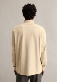 Beżowa koszula z długim rękawem, z wysokim kołnierzem i prostym krojem, wykonana z miękkiego materiału, charakteryzująca się gładką fakturą i minimalistycznym designem.