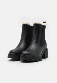 Bottines en cuir noir avec un talon épais et une semelle en caoutchouc texturé. Finition en fourrure sur le haut, avec des panneaux élastiques sur les côtés pour un enfilage facile.