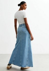Falda maxi de mezclilla en azul claro con cintura alta, bolsillos traseros y costuras verticales distintivas. Combinada con una camiseta blanca ajustada y zapatos.