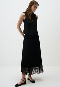 Gilet sans manches noir avec trois boutons, associé à une longue jupe en dentelle noire avec un motif texturé. Porté avec des ballerines noires simples.
