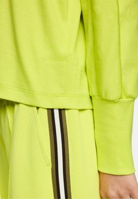 Neon-gelbes Sweatshirt mit Bündchen und Saum über passenden perforierten Shorts, die schwarze, weiße und olivgrüne vertikale Streifen entlang der Taschenkante aufweisen.
