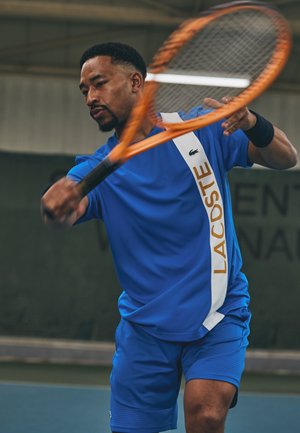 Giocatore di tennis con una maglia blu con una striscia bianca su cui è scritto "LACOSTE" in oro. Tiene in mano una racchetta arancione e colpisce una palla al coperto.