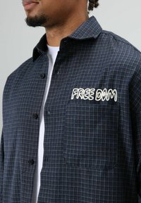 Blauw geruit overhemd met knopen van katoenen stof, met een borstzakje waarop witte geborduurde tekst "FREE DIM" staat, en een textuurpatroon detail.