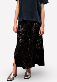Person in einem schwarzen, mit Blumenmotiven bedruckten Spitzensock, mit seitlichem Schlitz, einer dunkel gewaschenen, lockeren T-Shirt und metallischen silbernen Slippern, steht vor weißem Hintergrund.