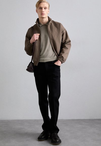 Ruskea teksturoitu pommik Jacket, vetoketjulla, vihreä polo-paita, mustat kapeat housut ja mustat nahkakengät. Malli pitää pientä ruskeaa laukkua.