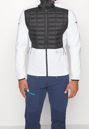 Mężczyzna w białej kurtce z czarnym pikowanym przodem, granatowych spodniach z kieszeniami na zamek, stojący z rękami swobodnie opuszczonymi.