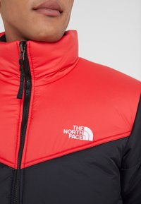 Giacca isolante rossa e nera con colletto alto, dotata di chiusura con zip e del logo bianco "The North Face" sul petto.