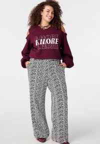 Sweatshirt bordeaux avec épaules découpées et texte "AMORE", associé à un pantalon large imprimé zèbre noir et blanc. Chaussures bordeaux brillantes.