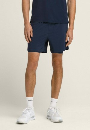 Mann trägt marineblaue Sportshorts, weiße Sportsocken mit Wilson-Logo und weiße Laufschuhe, steht vor einem einfarbigen Hintergrund.