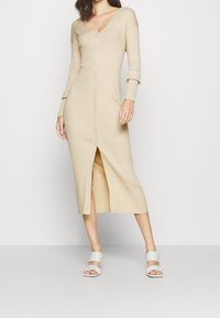 Beige ribbad stickad klänning med långa ärmar, V-ringning, knappstängning framtill och hög slits på låret. Matchas med vita sandaler med remmar.