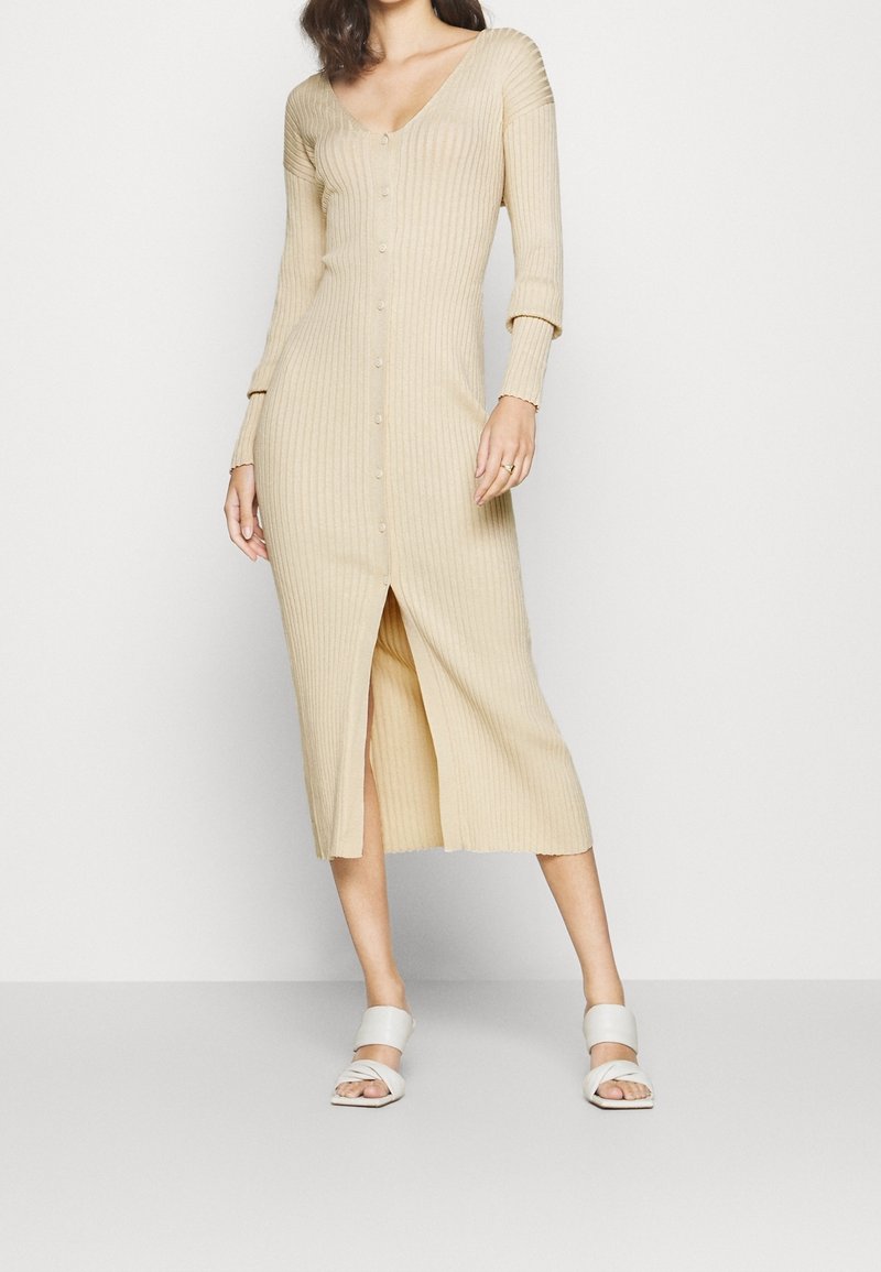 Beige ribbad stickad klänning med långa ärmar, V-ringning, knappstängning framtill och hög slits på låret. Matchas med vita sandaler med remmar.