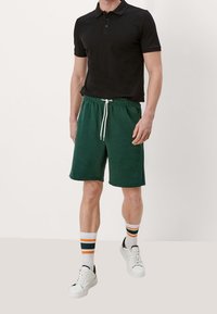 Shorts de cycliste verts avec une taille à cordon, des poches latérales et un logo brodé. Associés à un polo noir et des chaussettes rayées.