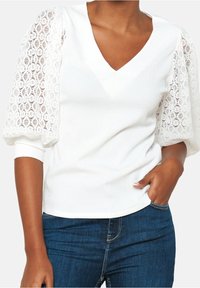 Biała bluzka z dekoltem w kształcie V, teksturowanym ciałem i bufiastymi rękawami z koronki w kwiatowy wzór. Połączona z niebieskimi dżinsami.