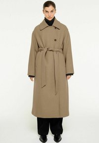 Lång, beige ullrock med en bred krage, enkel knappknäppning och bälte i midjan. Har vida ärmar och en rak passform.