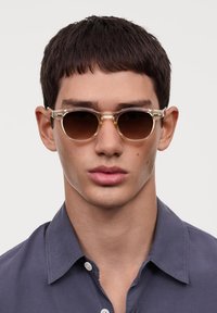 Ochelari de soare cu nuanță maro, cu montură din plastic transparent, având un design elegant și accente metalice. Purtați de o persoană cu părul scurt și închis la culoare.