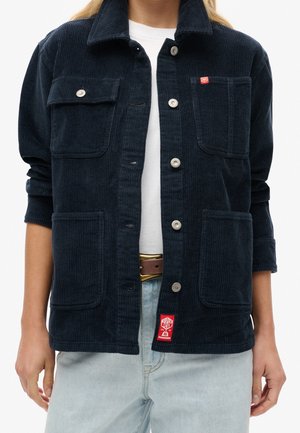 Woman wearing a navy blue corduroy jacket with four pockets and silver snap buttons over a white shirt and light blue jeans.