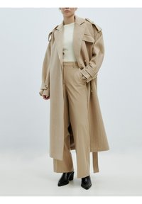 Beige übergroßer Trenchcoat mit Schulterklappen, ribbeligem cremefarbenen Oberteil und beigen gestreiften Hosen. Schwarze Stiefeletten runden das Outfit ab.