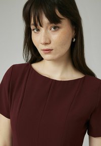 Robe bordeaux à manches courtes, décolleté arrondi et coutures verticales. Tissu lisse avec une coupe ajustée. Boucles d'oreilles en métal argenté.