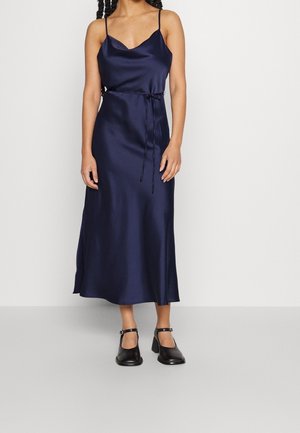 Vrouw draagt een marineblauwe satijnen midi slipjurk met dunne bandjes en een taillelint, gecombineerd met zwarte blokhak schoenen met enkelbandje.
