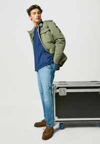 Chaqueta militar verde sobre una camiseta azul, combinada con jeans de un azul claro y zapatos de ante marrón. Caja de almacenamiento rodante negra al lado.
