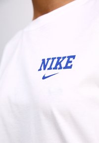 Bílá bavlněná tričko s výrazným modrým logem "NIKE" a menším modrým swoosh, kulatým výstřihem, hladkou texturou a krátkými rukávy.