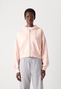 Hoodie cropped di colore rosa pastello, realizzato in morbido tessuto, con zip frontale, cappuccio con cordino e tasche laterali, abbinato a pantaloni della tuta grigi.