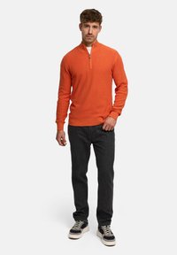 Orange strikket sweater med halv-zip krave, parret med mørke jeans og sorte sneakers med hvide såler og accenter.