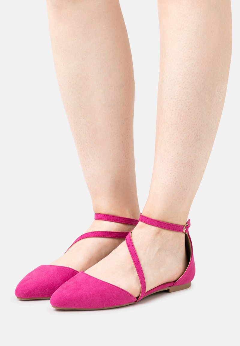 Pieds portant des chaussures plates pointues rose vif avec des brides croisées et autour de la cheville sur une peau claire et un fond blanc.