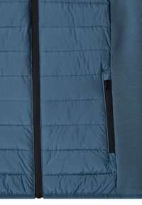 Section de veste en tissu matelassé bleu avec des coutures horizontales, avec une fermeture éclair verticale noire et un tissu bleu lisse sur le côté droit.