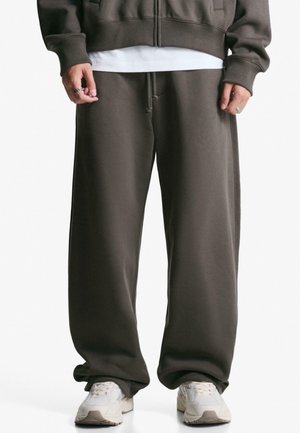 Pantaloni della tuta marrone scuro con vestibilità ampia, realizzati in tessuto morbido, caratterizzati da una vita elastica e tasche laterali. Abbinati a sneakers bianche.