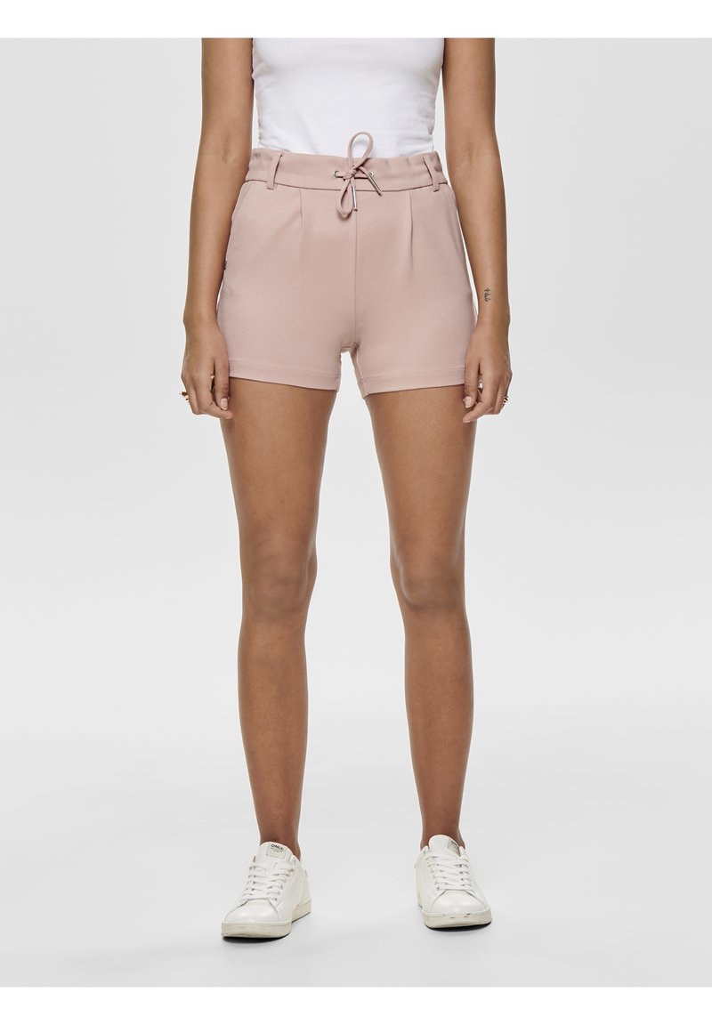 Woman wearing light pink high-waist shorts with a front bow tie, white tank top, and white sneakers, standing against a plain background.