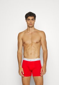 Rde boksarice iz bombaža z belim pasom, na katerem je napis "TOMMY HILFIGER", prikazujoče prilega dizajn in gladko teksturo.