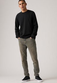 Black long-sleeve shirt, ribbed texture; faded gray jeans; black sneakers with white soles; model stands with hands in pockets.
