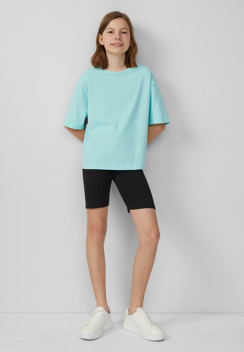 Lichtblauwe oversized t-shirt van katoen, gecombineerd met zwarte shorts. Het model draagt ook witte sneakers en staat tegen een effen achtergrond.